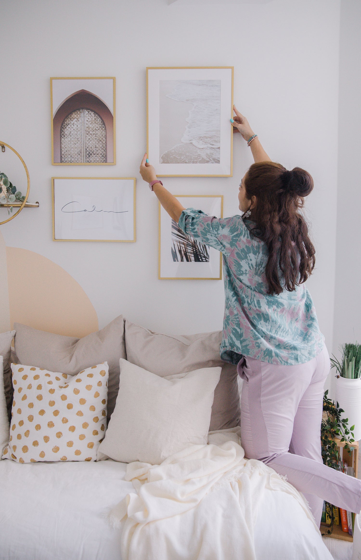 Woman adjusting framed artwork on white wall above beige sofa with patterned and plain pillows in cozy living room